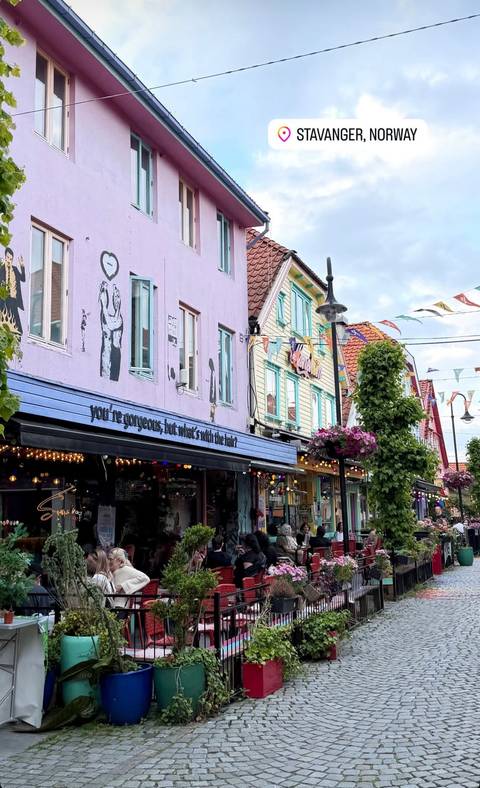 Colorful wooden shopfronts and pastel houses on a festive pedestrian street adorned with bunting and hanging flowers.