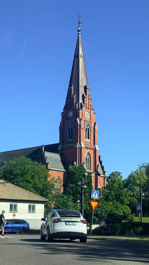 Tall red-brick neo-gothic church tower with clock faces rising above green trees and a vivid blue sky.