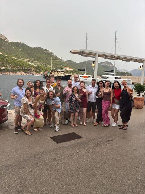 Well-dressed travelers toast with wine by a marina lined with yachts and green hills.
