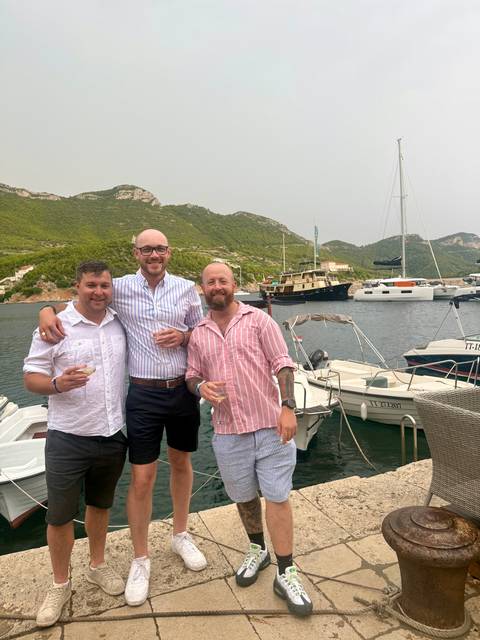 Three friends enjoy drinks on a quayside with yachts and forested hills in the background.