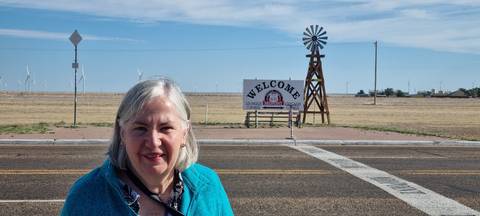 Traveller stands by a Route 66 midpoint welcome sign in a flat, windswept Texas landscape.