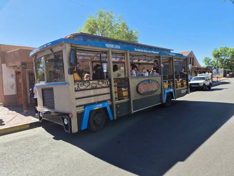 Open-air vintage trolley packed with tourists drives through a sunny Southwestern town.