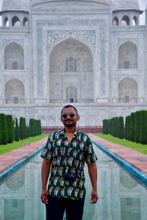 Visitor poses with sunglasses before the marble façade of the Taj Mahal, framed by gardens and cypress trees.