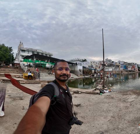 Selfie with stepped ghats and a calm lake under a cloudy sky in an Indian town setting.