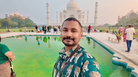 Traveller takes a selfie beside a green reflecting pool with the Taj Mahal in soft morning haze.