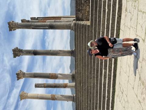 Smiling couple standing before ancient stone columns and steps at the Roman ruins of Volubilis.