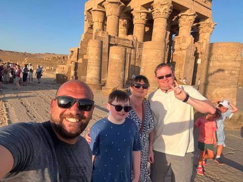 A smiling family takes a selfie in front of sun-lit sandstone columns at Kom Ombo Temple.