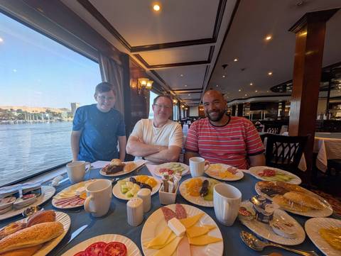 Three travelers enjoy a hearty breakfast spread on a Nile cruise ship with river views through large windows.