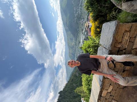 Traveler sits on a stone wall overlooking a lush Bhutanese valley ringed by green mountains and scattered villages.