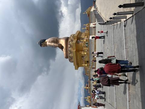 Visitors walk toward a colossal golden seated Buddha statue under dramatic clouds on a hillside plaza.