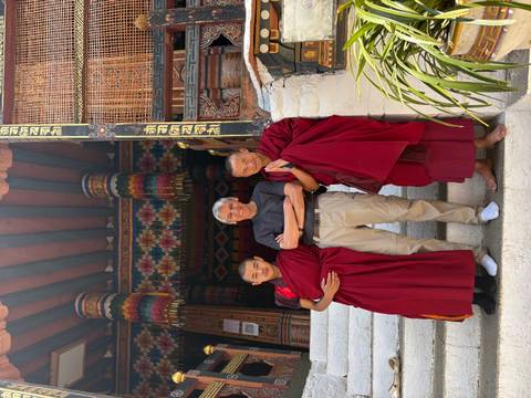 A traveler poses arm-in-arm with two monks in crimson robes on monastery steps decorated with colorful woodwork.