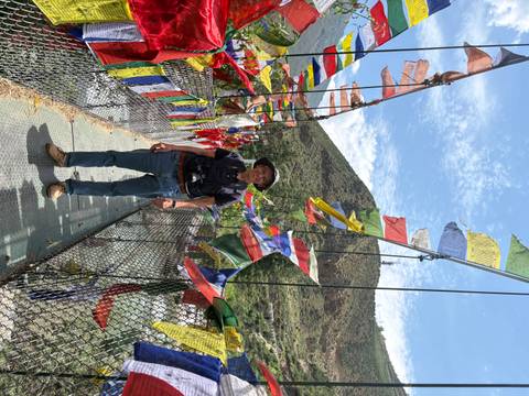 Traveler stands on a suspension bridge festooned with colorful prayer flags spanning a river gorge.