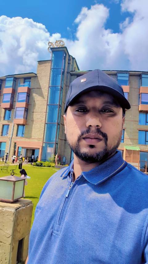 Close-up selfie of a man in a cap with a modern hotel facade and lawn behind him.