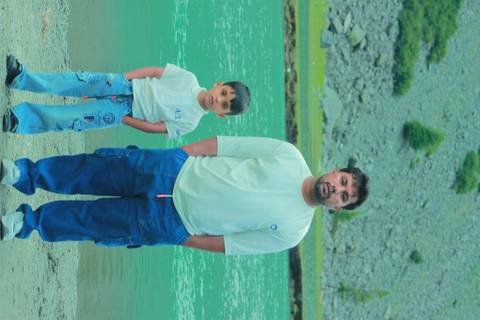 Man and child stand on the bank of a glacial green river with rocky hills behind.