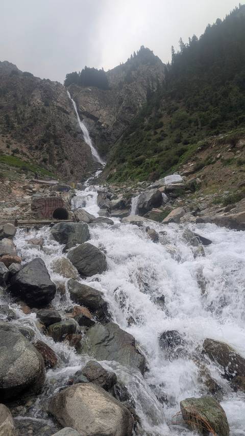 White water tumbles over boulders in a steep mountain ravine with a distant waterfall.