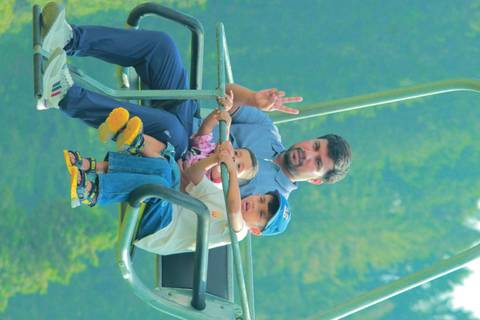 Father and two children ride an open chairlift above green forest, father flashing a peace sign.