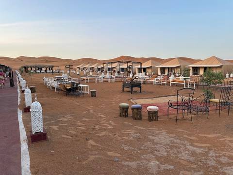 A desert camp with canvas tents, metal lanterns, and fire pits sits on reddish sand dunes at dusk.