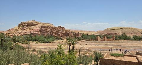 View across a dry riverbed towards the earthen citadel of Ait Benhaddou framed by desert hills and scattered greenery under a clear blue sky.
