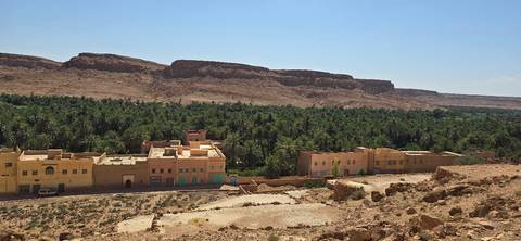 Line of pastel buildings fronting a lush palm oasis backed by rugged arid cliffs under a bright sky.