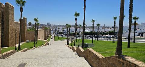 Stone ramp lined with palm trees leading from city walls down to a modern avenue and skyline in the distance.