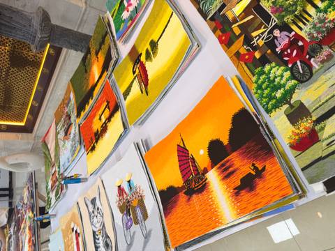 Stacks of vibrant hand-embroidered art pieces depicting Vietnamese countryside scenes on a market stall.