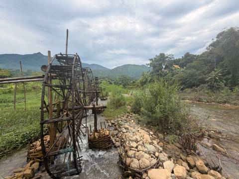 Traditional bamboo water wheels line a small rocky stream with green mountains in the background under a cloudy sky.