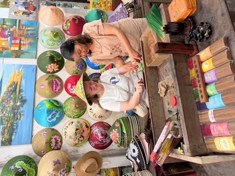 A traveller and a local craftswoman prepare colorful incense sticks amid vibrant conical hats and art pieces.