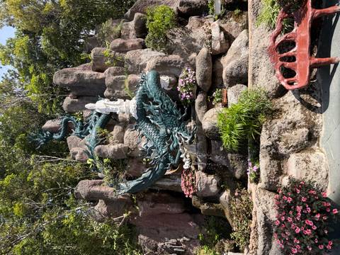 An elaborate blue-green dragon sculpture curls around rocks with a white deity statue above, surrounded by flowers and greenery.