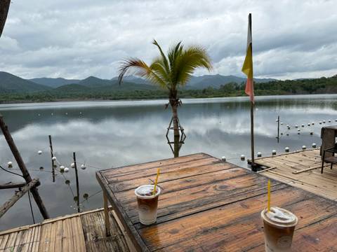 Serene lakeside café table with iced coffees, overlooking calm water, palm tree, and mountain backdrop under gray skies