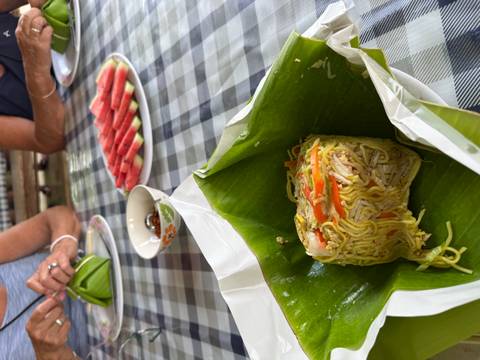 Portion of fried noodles wrapped in banana leaf with sliced watermelon in background on a checkered table
