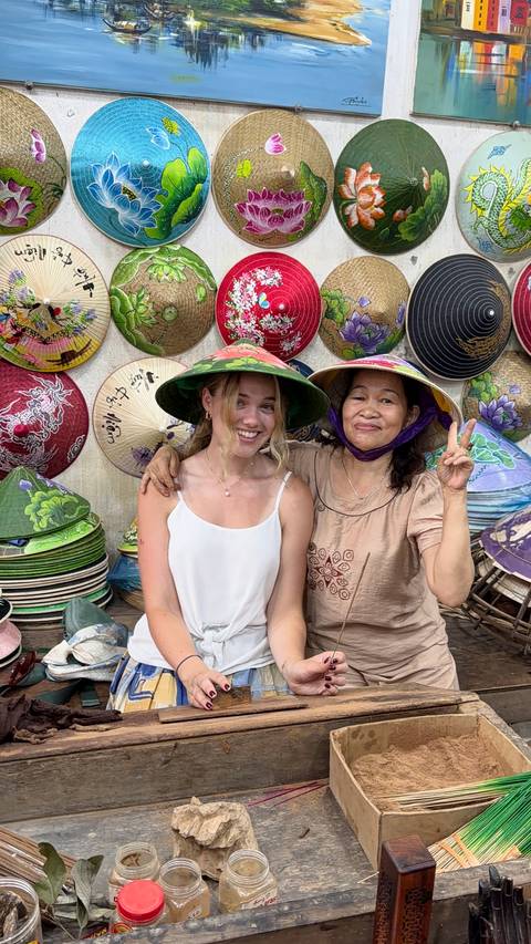 Smiling traveler posing with local vendor, both wearing traditional conical hats in a colorful hat shop