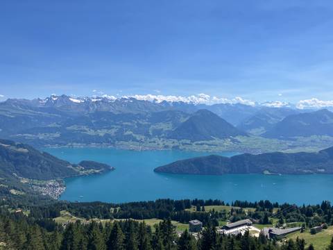 Panoramic high-altitude view of a turquoise lake encircled by green valleys and snow-dusted Alps