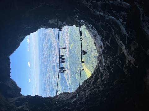 Rocky cave opening framing a valley far below, with via ferrata cables and carabiners in the foreground