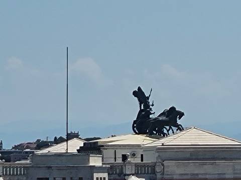 Silhouette of a chariot statue atop a classical building roof against a bright midday sky in Rome.
