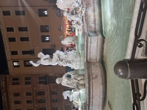 Night-lit marble statues in a Roman fountain surrounded by glowing windowed buildings.