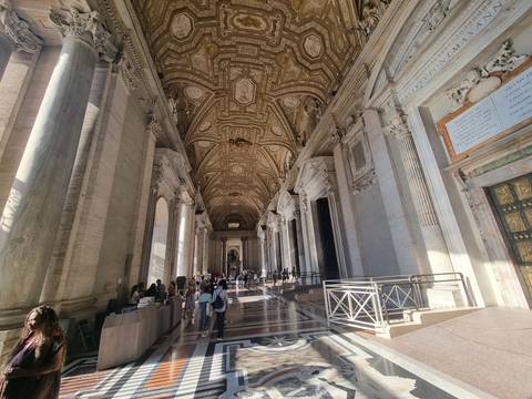Grand marble corridor inside St. Peter’s Basilica bathed in warm afternoon light with visitors exploring.
