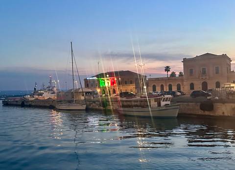 Harbor scene at dusk with boats moored beside historic buildings lit by colorful lights.