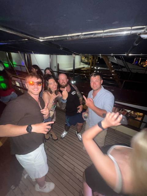 Friends party on a yacht deck at night, flashing hand signs toward the camera.