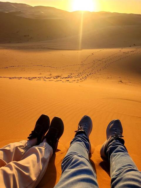 View from above of golden sand with footprints, framed by three pairs of resting feet.