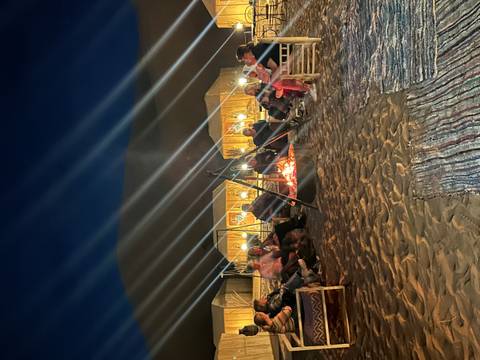 Nighttime campfire circle on desert sand with tents behind and beams of light streaking overhead.