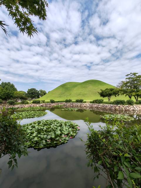 Lily-fringed pond reflects twin grassy burial mounds beneath scattered clouds in Gyeongju.