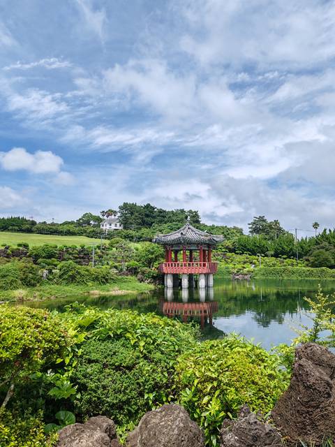 Traditional red pavilion stands on stilts over a reflective pond in a manicured Jeju garden.