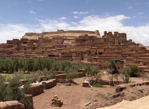 Earth-toned kasbah town stacked on a hilltop with green oasis foreground under patchy clouds.