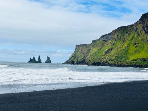 Black-sand beach with crashing waves, green cliffs and dramatic sea-stack formations.