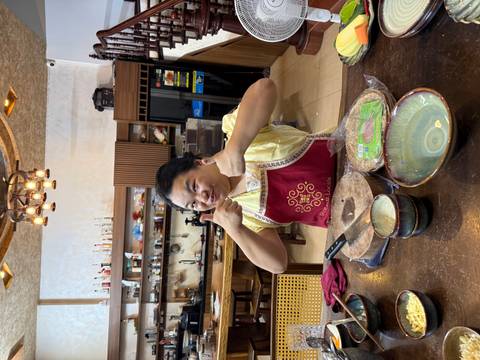 Smiling cooking instructor wearing an apron gives two thumbs up inside a cozy kitchen classroom.