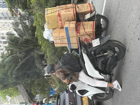 Motorbike rider with helmet balances stacks of cardboard boxes tied to the back in busy city traffic.