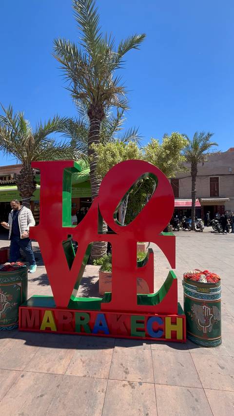 Large red and green LOVE sculpture decorates a sunny market plaza with passersby and shops.