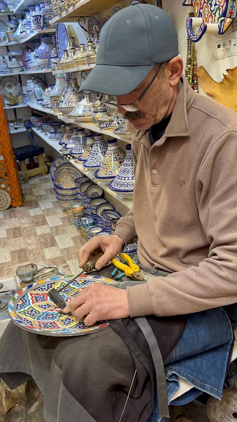 Artisan works on metal against shelves of colorful Moroccan pottery in a shop.