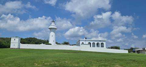 White lighthouse and adjoining building on a wide green lawn beneath a bright blue sky