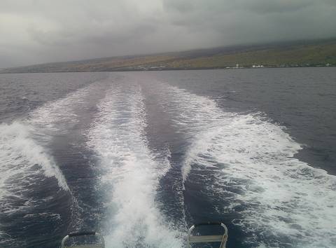 Boat wake trails across deep blue ocean with distant volcanic coastline under grey sky